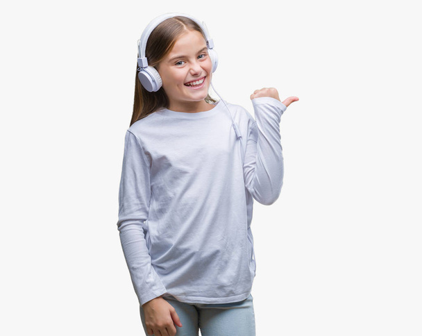 Young beautiful girl wearing headphones listening to music over isolated background smiling with happy face looking and pointing to the side with thumb up. - Photo, Image