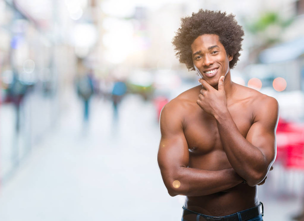 Hombre afroamericano sin camisa mostrando cuerpo desnudo sobre fondo aislado mirando confiado a la cámara con sonrisa con los brazos cruzados y la mano levantada en la barbilla. Pensando positivamente
. - Foto, imagen