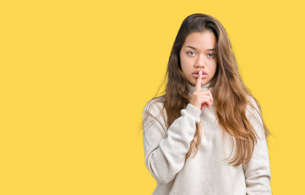 若い美しいブルネット女性の唇に指で静かにするように求めて孤立の背景にタートルネックのセーターを着ています。沈黙と秘密の概念. - 写真・画像