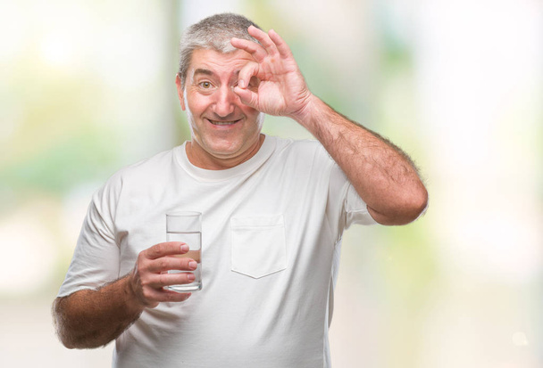 Hombre mayor guapo bebiendo vaso de agua sobre fondo aislado con la cara feliz sonriendo haciendo signo bien con la mano en el ojo mirando a través de los dedos
 - Foto, imagen