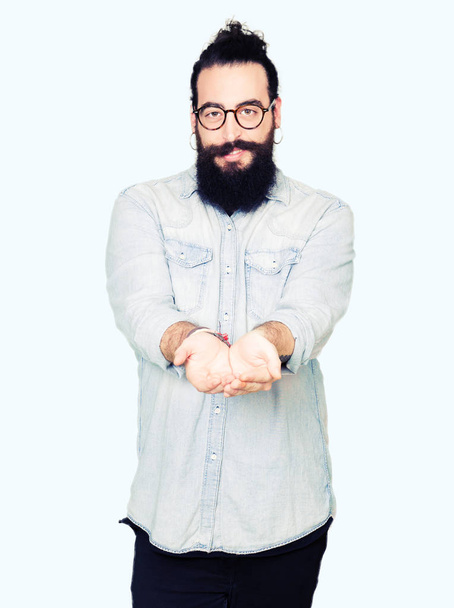 Homem jovem hipster com cabelos longos e barba usando óculos Sorrindo com as mãos palmas juntas recebendo ou dando gesto. Seguro e proteção
 - Foto, Imagem