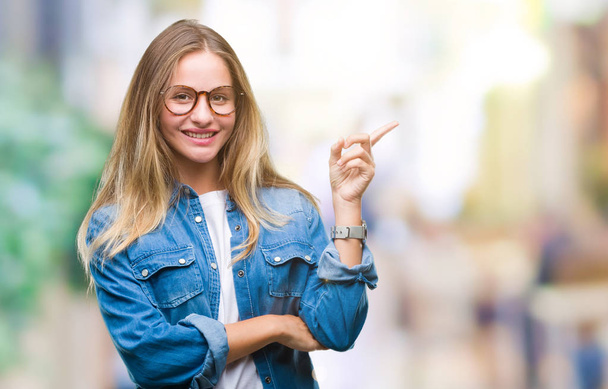 junge schöne blonde Frau trägt eine Brille über isoliertem Hintergrund mit einem breiten Lächeln im Gesicht und zeigt mit der Hand und dem Finger zur Seite in die Kamera. - Foto, Bild