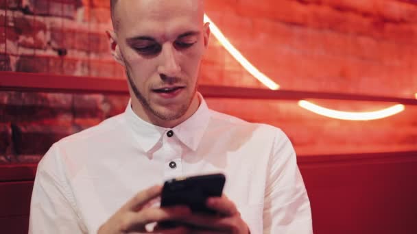Young Attractive Man Using Smartphone at Evening. He standing in a bar or restaurant near neon signage. Communication, rest, chatting, travel concept. Close up - Filmagem, Vídeo