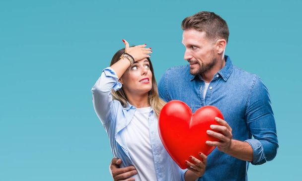 Young couple in love holding red heart over isolated background stressed with hand on head, shocked with shame and surprise face, angry and frustrated. Fear and upset for mistake. - Fotografie, Obrázek