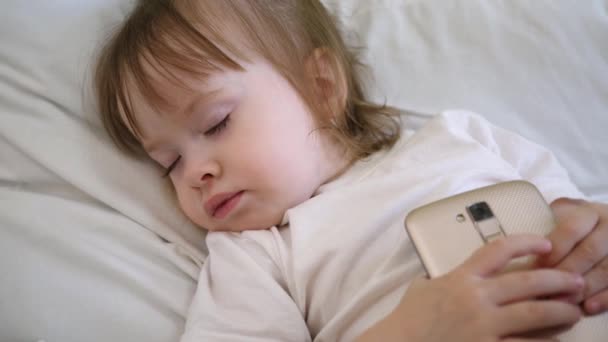 dítě leží na polštáři a držení tabletu. Roztomilé dítě spí v posteli s smartphone. - Záběry, video