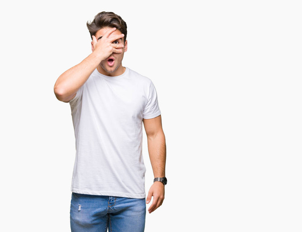 Joven hombre guapo vistiendo una camiseta blanca sobre un fondo aislado mirando con la mano a través de los dedos con expresión avergonzada
. - Foto, Imagen