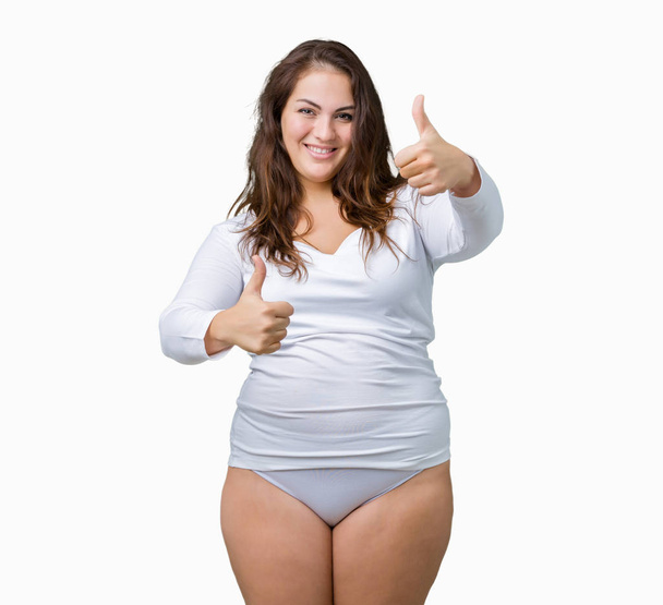 Beautiful plus size young overwight woman wearing white underwear over isolated background approving doing positive gesture with hand, thumbs up smiling and happy for success. Looking at the camera, winner gesture. - Photo, Image