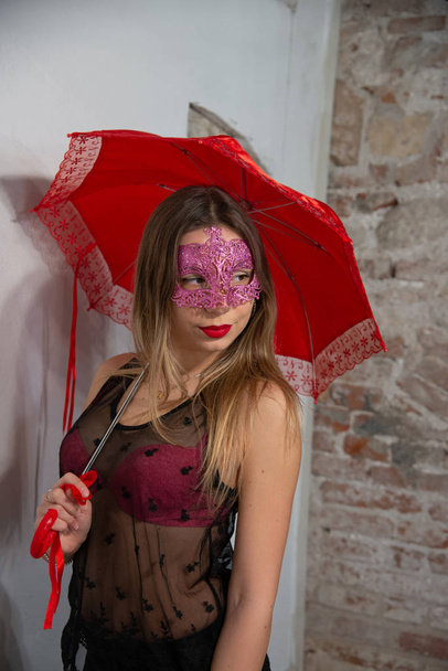 ベネチア カーニバル赤マスク、赤い傘と、美しい若い女性の肖像画。完璧なメイクで透明のタンクトップとピンクのブラジャーのセクシーなブロンドのグラマー ガール。マスクのパーティーのための魅力的な写真. - 写真・画像