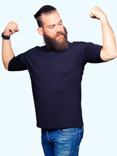 Joven hombre rubio con camiseta casual mostrando los músculos de los brazos sonriendo orgulloso. Concepto Fitness
. - Foto, imagen