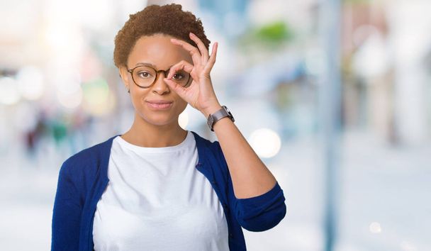 Jonge mooie Afrikaanse Amerikaanse vrouw bril over geïsoleerde achtergrond doen ok gebaar met hand glimlachen, oog kijken door vingers met blij gezicht. - Foto, afbeelding