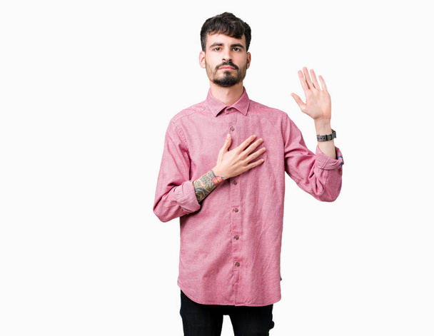 Молодой красивый мужчина в розовой рубашке на изолированном фоне, ругаясь с рукой на груди и открытой ладони, давая клятву верности
 - Фото, изображение