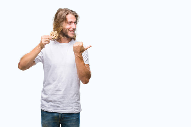Young handsome man with long hair eating chocolate cooky over isolated background pointing and showing with thumb up to the side with happy face smiling - Photo, Image