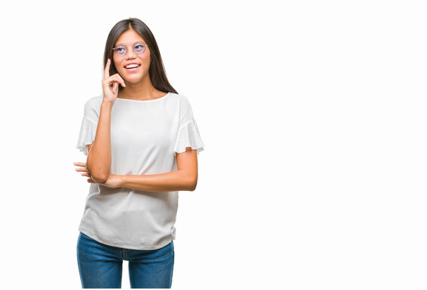 junge asiatische Frau mit Brille vor isoliertem Hintergrund, die Hand am Kinn, nachdenklich, fragend. Lächeln mit nachdenklichem Gesicht. Zweifelhaftes Konzept. - Foto, Bild