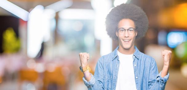 Americký mladík s afro vlasy nosí brýle slaví překvapen a ohromen pro úspěch s náručí vystouplý a otevřené oči. Vítěz koncept. - Fotografie, Obrázek