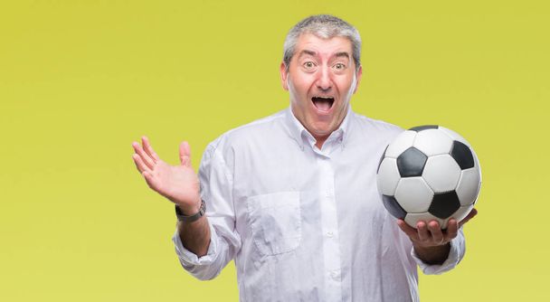 Handsome senior man holding soccer football ball over isolated background very happy and excited, winner expression celebrating victory screaming with big smile and raised hands - Фото, изображение