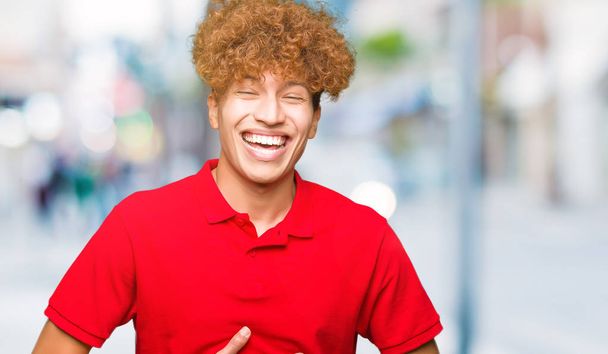 Молодой красивый мужчина с афроволосами в красной футболке улыбается и громко смеется, потому что смешная сумасшедшая шутка. Счастливое выражение
. - Фото, изображение