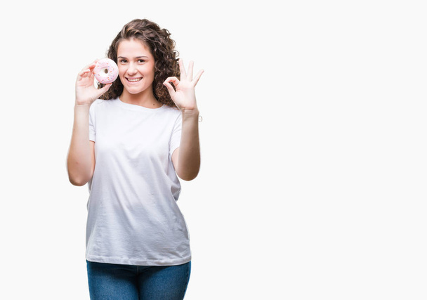 Young brunette girl eating donut over isolated background doing ok sign with fingers, excellent symbol - Foto, immagini