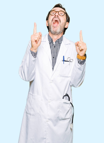 Середнього віку лікарі чоловіки в медичному пальто здивовані і здивовані, дивлячись вгору і вказуючи пальцями і підняті руки
. - Фото, зображення