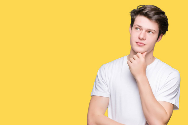 Young handsome man wearing casual white t-shirt over isolated background with hand on chin thinking about question, pensive expression. Smiling with thoughtful face. Doubt concept. - Photo, Image