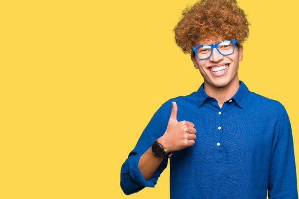 Homem bonito jovem com cabelo afro vestindo óculos azuis fazendo polegares felizes até gesto com a mão. Aprovando expressão olhando para a câmera com mostrando sucesso
. - Foto, Imagem