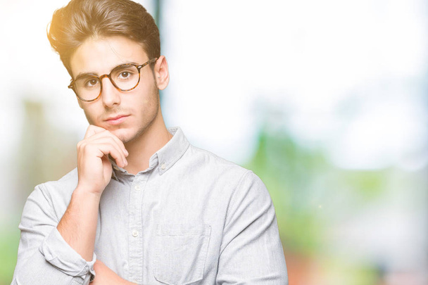 Young handsome man wearing glasses over isolated background with hand on chin thinking about question, pensive expression. Smiling with thoughtful face. Doubt concept. - Photo, Image