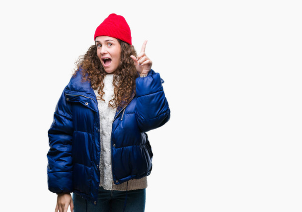Beautiful young brunette curly hair girl wearing winter coat, wool cap and sweater over isolated background pointing finger up with successful idea. Exited and happy. Number one. - Photo, Image