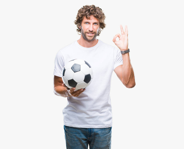 Όμορφος άνδρας Ισπανόφωνος μοντέλο κρατώντας την μπάλα ποδοσφαίρου ποδοσφαίρου πάνω από απομονωμένες φόντο κάνει εντάξει πινακίδα με τα δάχτυλα, εξαιρετική σύμβολο - Φωτογραφία, εικόνα