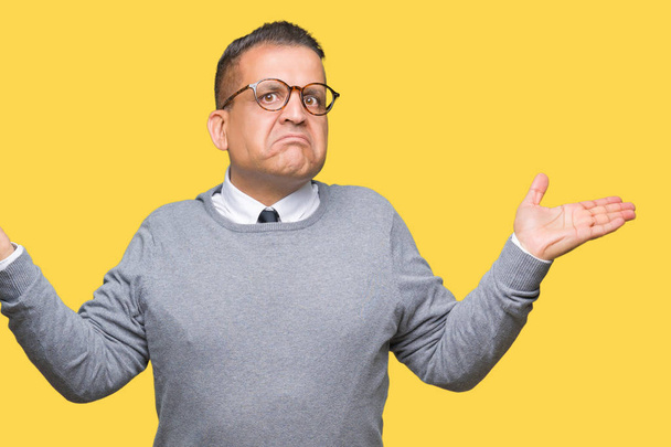 Middle age bussines arab man wearing glasses over isolated background clueless and confused expression with arms and hands raised. Doubt concept. - Photo, Image
