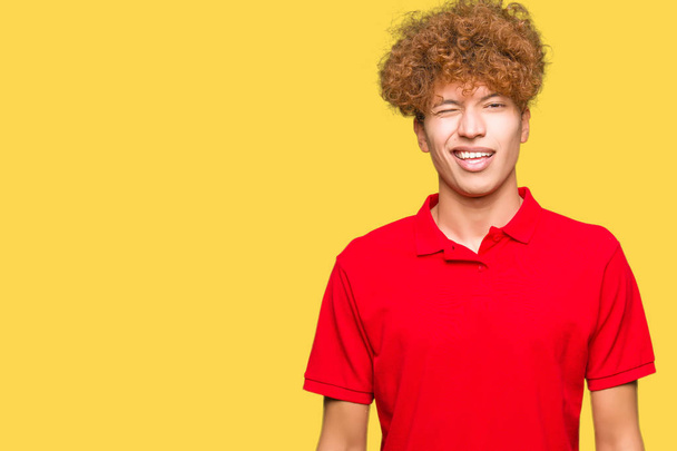 Joven hombre guapo con pelo afro vistiendo camiseta roja guiñando el ojo mirando a la cámara con expresión sexy, cara alegre y feliz
. - Foto, Imagen
