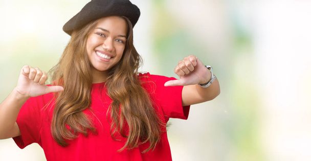 junge schöne brünette Modefrau trägt rotes T-Shirt und schwarzes Baskenmütze über isoliertem Hintergrund und sieht selbstbewusst mit einem Lächeln auf dem Gesicht aus, zeigt mit den Fingern stolz und glücklich. - Foto, Bild