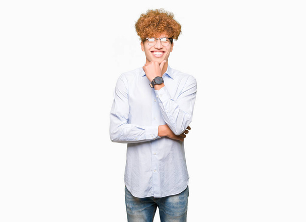 Young handsome business man with afro wearing glasses looking confident at the camera with smile with crossed arms and hand raised on chin. Thinking positive. - Photo, Image
