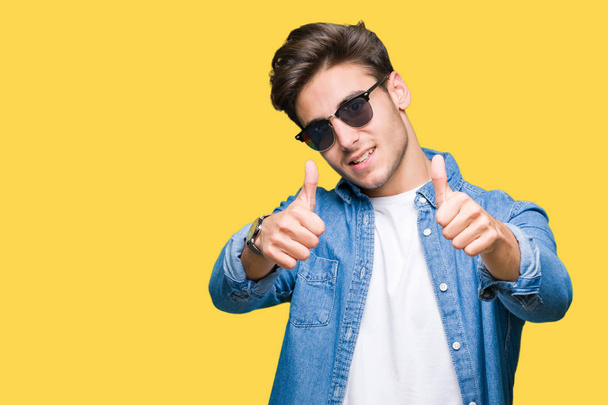 Young handsome man wearing sunglasses over isolated background approving doing positive gesture with hand, thumbs up smiling and happy for success. Looking at the camera, winner gesture. - Photo, Image