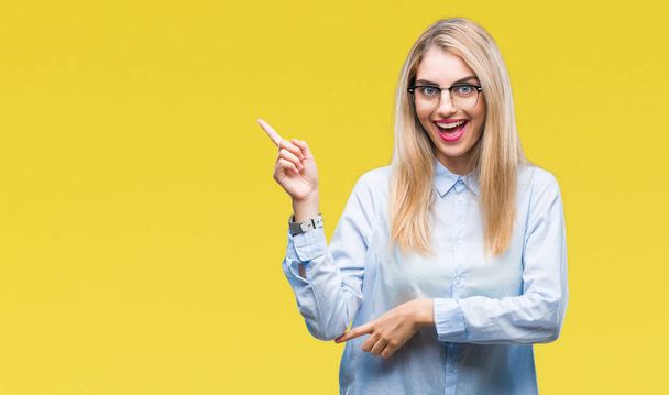 Young beautiful blonde business woman wearing glasses over isolated background with a big smile on face, pointing with hand and finger to the side looking at the camera. - Photo, Image