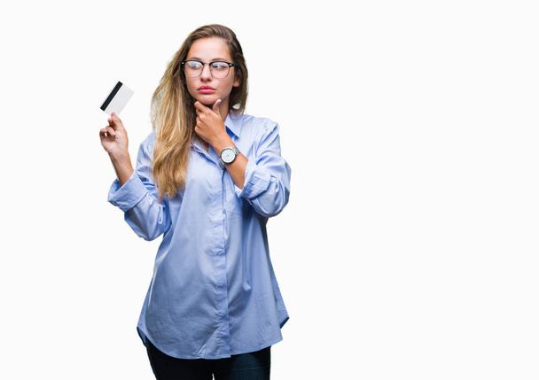 Young beautiful blonde woman holding credit card over isolated background serious face thinking about question, very confused idea - Foto, afbeelding