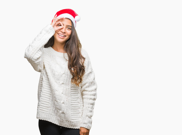 Mujer árabe joven con sombrero de Navidad sobre fondo aislado haciendo buen gesto con la mano sonriente, ojos mirando a través de los dedos con la cara feliz
. - Foto, imagen