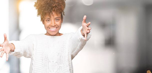 Красива молода жінка афроамериканець, носити светр на зиму над ізольованих фону, дивлячись на камеру, посміхаючись з розкритими обіймами обійми. Веселий вираз всеосяжне щастя. - Фото, зображення
