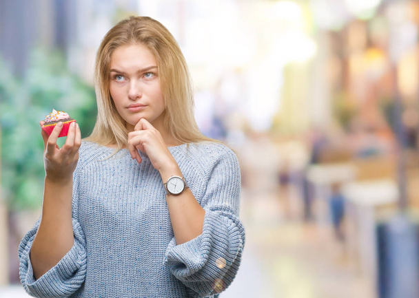 Mujer caucásica joven comiendo magdalena dulce sobre fondo aislado cara seria pensando en la pregunta, idea muy confusa
 - Foto, imagen