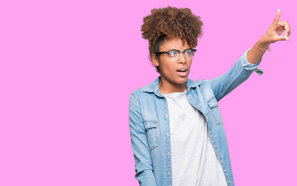 Bella giovane donna afroamericana che indossa occhiali su sfondo isolato Indicazione con il dito sorpreso avanti, bocca aperta espressione stupita, qualcosa di fronte
 - Foto, immagini