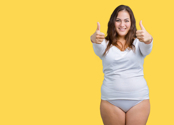 Beautiful plus size young overwight woman wearing white underwear over isolated background doing happy thumbs up gesture with hand. Approving expression looking at the camera showing success. - Photo, Image