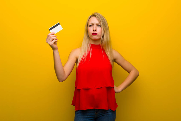 Jong meisje met rode jurk over gele muur nemen een creditcard zonder geld - Foto, afbeelding
