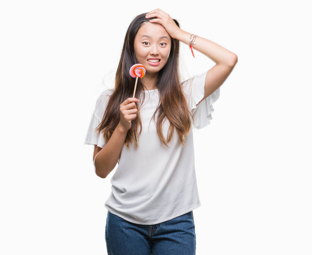 Mujer asiática joven comiendo caramelos de piruleta sobre fondo aislado estresado con la mano en la cabeza, conmocionado con la vergüenza y la cara sorpresa, enojado y frustrado. Miedo y malestar por error
. - Foto, imagen