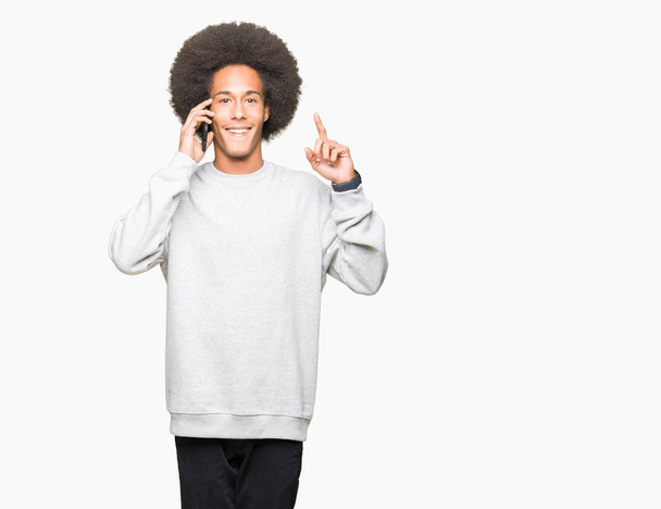 Nuori afrikkalainen amerikkalainen mies, jolla on afro-hiukset, puhuu älypuhelimella yllättynyt ideasta tai kysymyksestä, joka osoittaa sormella onnellisia kasvoja, numero yksi
 - Valokuva, kuva