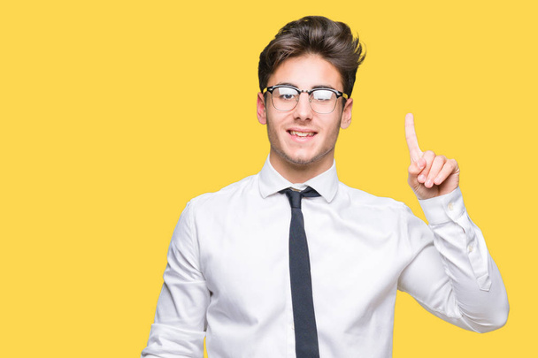 Jeune homme d'affaires portant des lunettes sur fond isolé montrant et pointant vers le haut avec le doigt numéro un tout en souriant confiant et heureux
. - Photo, image