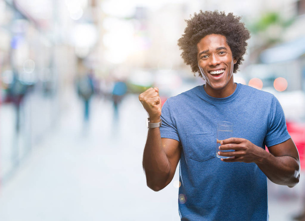 Afro americký muž pít sklenici vody nad izolované pozadí křičí hrdá a slaví vítězství a úspěch, velmi nadšený, fandění emoce - Fotografie, Obrázek