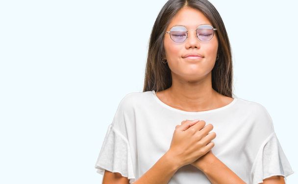 Szemüveg viselése alatt elszigetelt háttér, mosolygós, fiatal ázsiai nő kezét a mellkas csukott szemmel és hálás gesztus arcán. Egészségügyi koncepció. - Fotó, kép