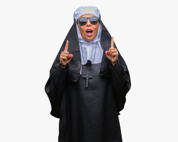 Orta yaş üst düzey Katolik Rahibe kadın izole arka plan üzerinde güneş gözlüğü takıyor hayret ve yukarıya bakıp parmakları ile işaret şaşırttı ve silah kaldırdı. - Fotoğraf, Görsel