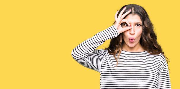 Young beautiful woman wearing stripes sweater doing ok gesture shocked with surprised face, eye looking through fingers. Unbelieving expression. - Photo, Image