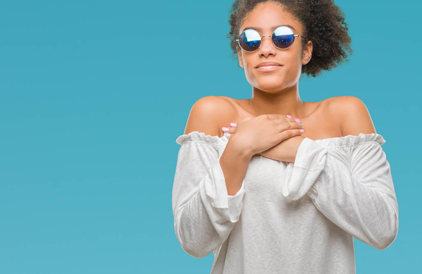 Молодих афро американської жінкою, носити сонцезахисні окуляри над ізольованих фон посміхається з руки на грудях з закритими очима і вдячний жест на обличчі. Концепція здоров'я. - Фото, зображення