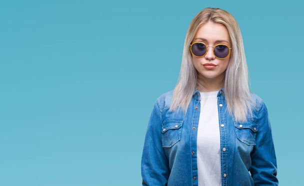 Young blonde woman wearing sunglasses over isolated background with serious expression on face. Simple and natural looking at the camera. - Photo, Image