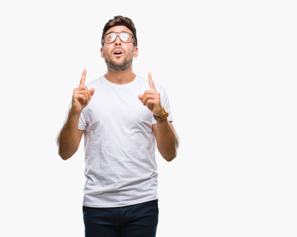 Young handsome man wearing glasses over isolated background amazed and surprised looking up and pointing with fingers and raised arms. - Photo, Image